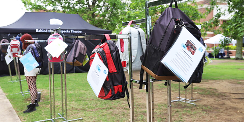 Rows of backpacks with notes on them hanging on racks along sidewalk.