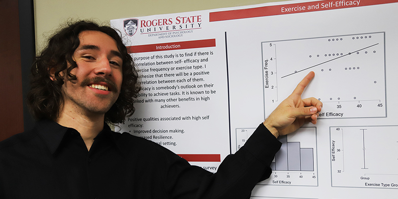 A male students smiles and points at his research poster.