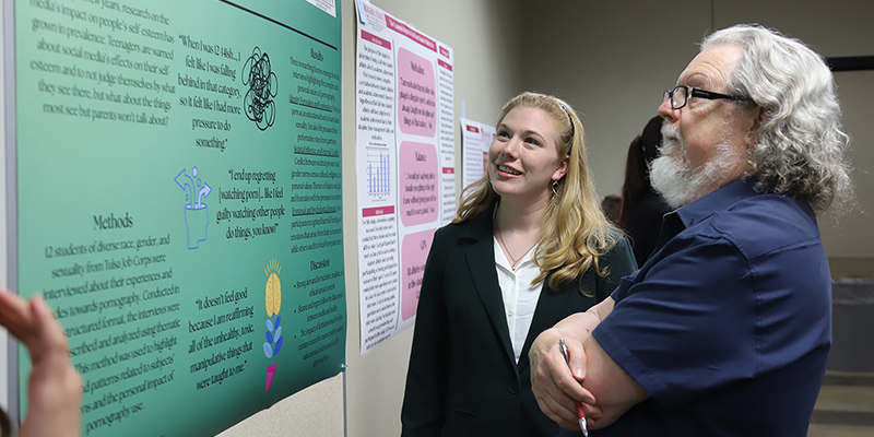 A female students disucses her research poster with a male professor.