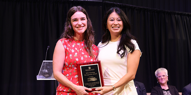 A smiling professor stands next to a smiling graduate