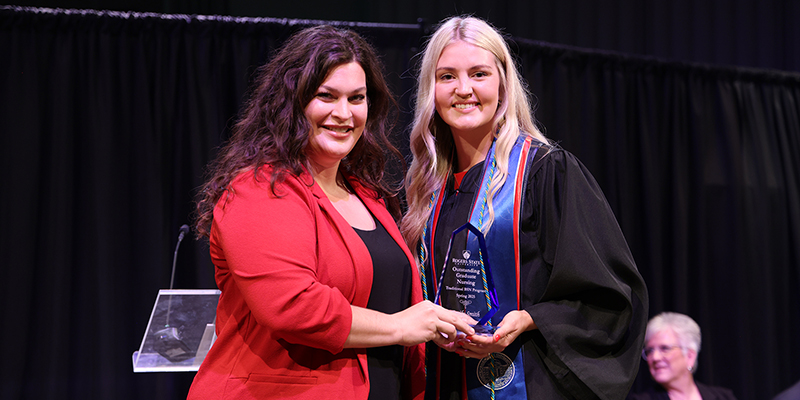 A smiling professor stands next to a smiling graduate