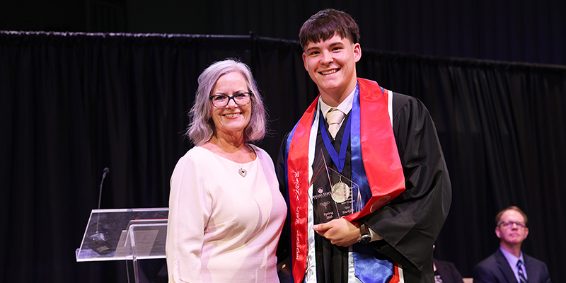 A smiling professor stands next to a smiling graduate