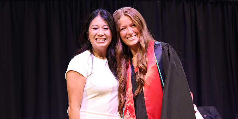 A smiling professor stands next to a smiling graduate
