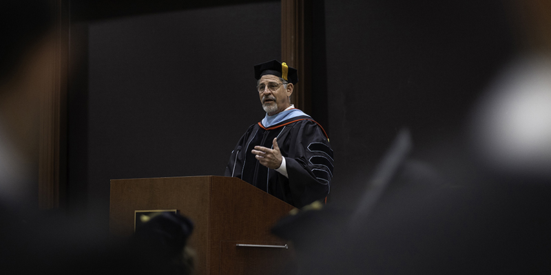 A man speaks at a podium while wearing academic regalia.