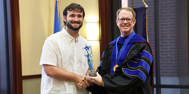 A smiling professor to the right present an award to a male student on the left.