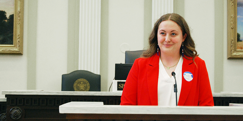 A young woman in a red blazer stands at a podium