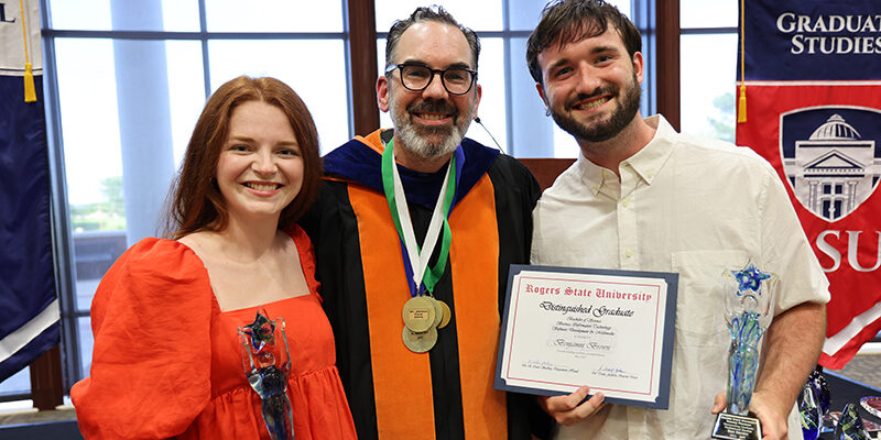 A smiling female student on the left is joined by a professor in the center and a male student on the right, all posing for a photo.