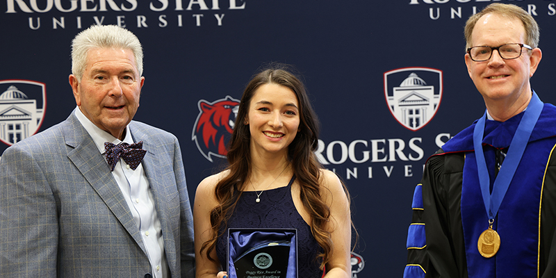 A smiling female student stands between two smiling men, as she holds and award trophy.