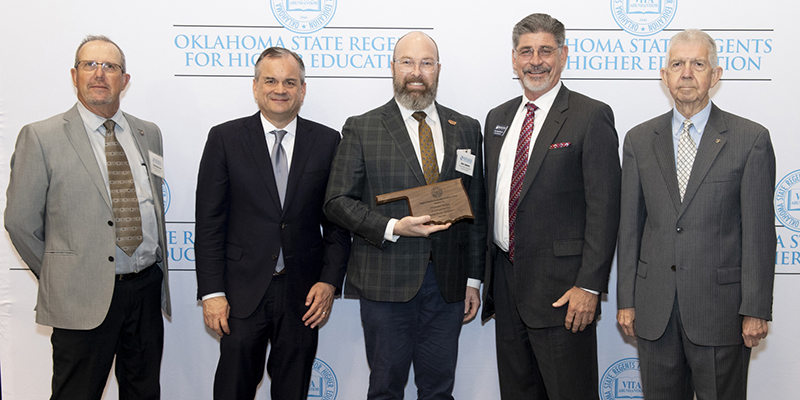 Five men pose for a photo with one in the center holding an award.