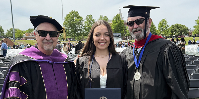 Two male professors stand on either side of a female student, all three in commencement robes