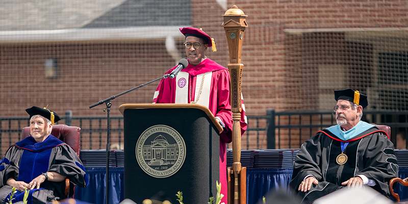A man in red and white academic robes speaks while standing at a podium.