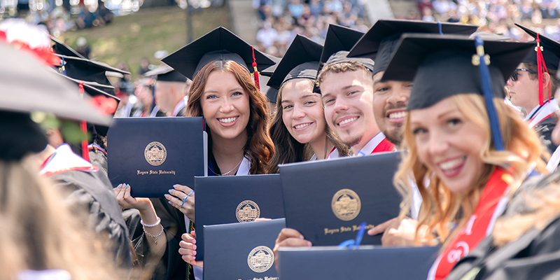 A group of graduates smile and hold their diploma covers up as they pose for a photo.