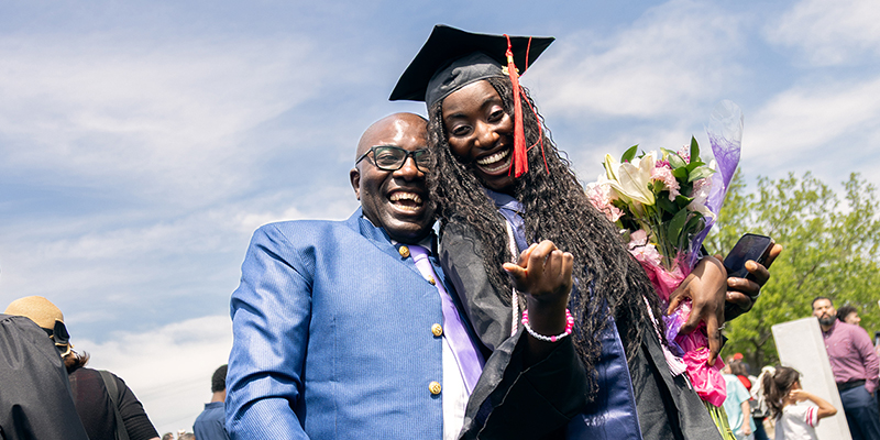 An adult male in a suit celebrates with an adult female in a graduation robe.