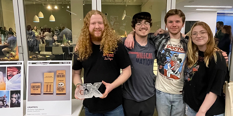 Four young adults stand smiling and posing with a photo, including one holding an award.