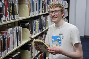 A man wearing glasses, standing in a library while holding an open boo