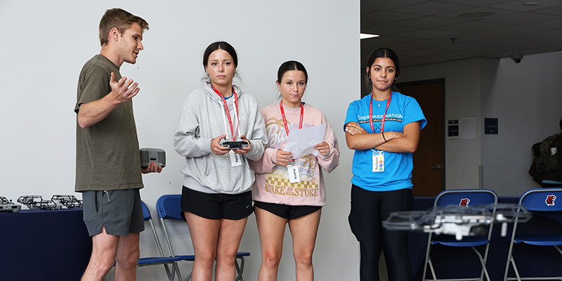 A male RSU student and three female high school students look at a flying drone